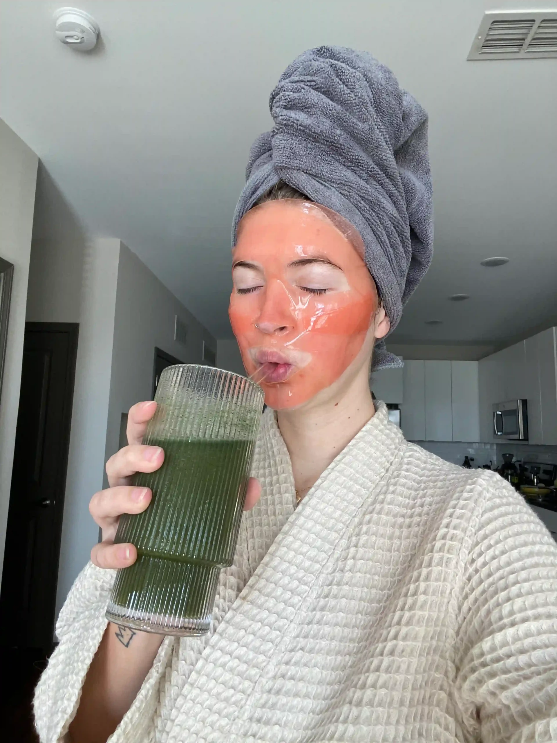 Mujer con el cabello envuelto en una toalla y mascarilla facial disfrutando de su rutina matutina de autocuidado