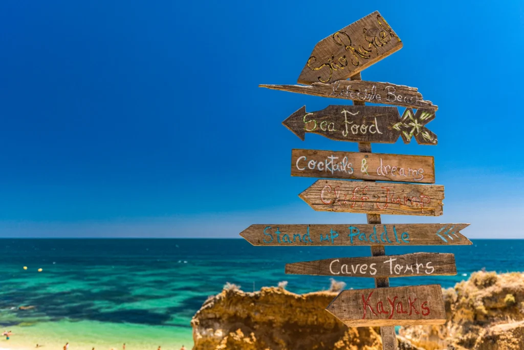 Señal de madera frente al mar con planes de vacaciones como playa, comida, cocktails, paddle, cuevas y kayak para adaptar los outfits al destino.