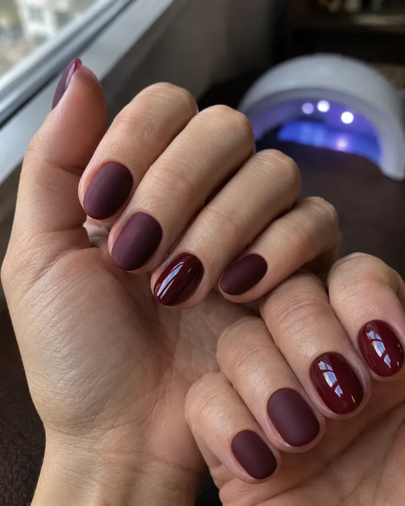 Uñas ovaladas cortas en burdeos combinando acabado mate y brillante con lámpara LED al fondo